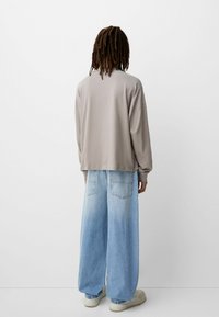 Svijetlo siva majica dugih rukava, ležernog kroja, uparena s širokim svijetlo plavim trapericama. Cipele su glomazne kremaste tenisice. Minimalistička pozadina.