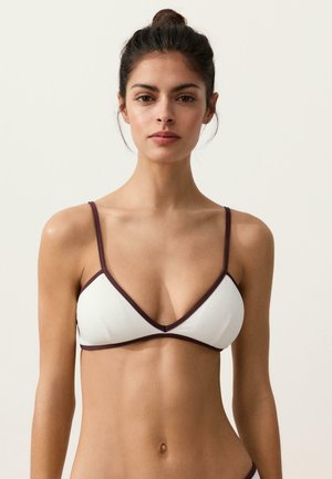 Tânără cu păr închis la culoare prins într-un coc, purtând un costum de baie alb cu cupe triunghiulare și margini închise la culoare, stând pe un fundal simplu, deschis la culoare.