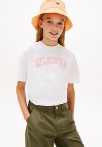 Λευκό κοντομάνικο μπλουζάκι με ροζ κείμενο "HILFIGER", συνδυασμένο με ελιά παντελόνια. Ένα ανοιχτό πορτοκαλί καπέλο τύπου bucket ολοκληρώνει την εμφάνιση.