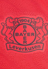 Tela roja con diseño texturizado que presenta un logotipo redondo negro de Bayer Leverkusen, flanqueado por dos leones, y el año 1904 mostrado de manera destacada.