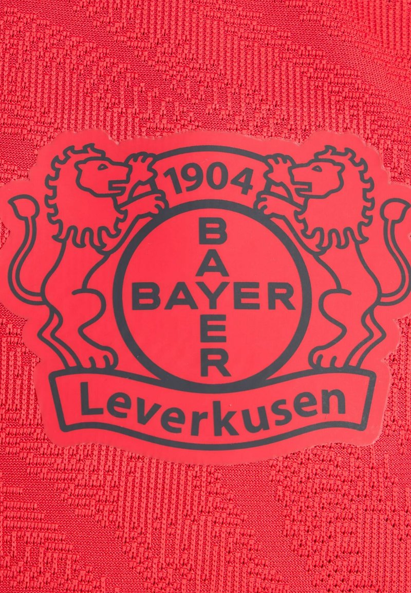 Roter Stoff mit strukturiertem Design zeigt ein rundes schwarzes Logo von Bayer Leverkusen, flankiert von zwei Löwen, und das Jahr 1904 wird deutlich angezeigt.