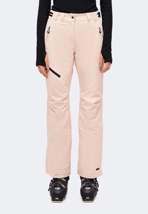 Pantalon de ski rose clair avec des fermetures éclair noires porté avec des chaussures de ski noires et un haut noir à manches longues, vu de la taille aux pieds.