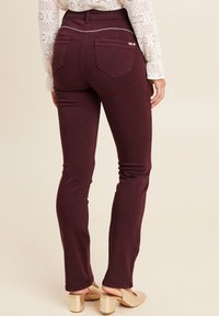Jeans slim fit maroon avec poches arrière et une seule couture décorative à la taille. Portés avec des chaussures à talons carrés de couleur claire.
