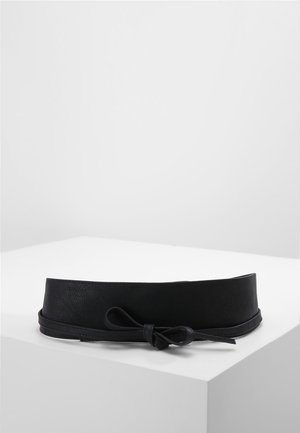 Szeroki czarny pasek z wąskim paskiem zawiązanym na kokardę na białej powierzchni na tle jednolitego jasnoszarego tła.