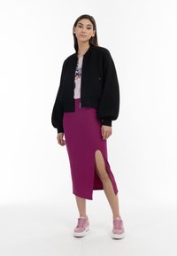 Zwarte cropped bomberjack, roze graphic t-shirt en een aansluitende magenta midi-rok met een split aan de zijkant, gecombineerd met roze sneakers.