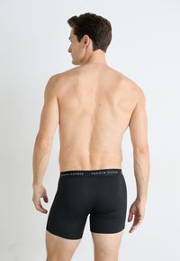 Schwarze Boxer-Brief aus Stretchstoff mit einem elastischen Bund, der den Text und das Logo "TOMMY HILFIGER" trägt. Modell von hinten betrachtet.