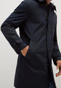 Laivastonsininen vedenpitävä trenssitakki suoralla leikkauksella, sivutaskuilla, napitettavilla hihansuihilla ja napilla suljettavalla edessä. Sileä pinta.