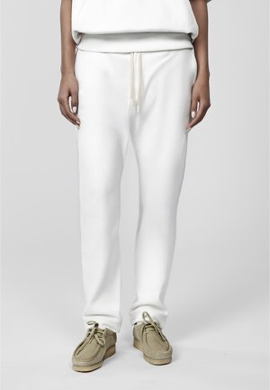 Personne portant un pantalon de survêtement blanc à cordon et des chaussures à lacets en daim beige, debout sur un fond gris clair uni.