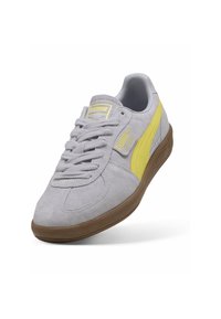 Baskets Puma en daim gris clair avec une bande latérale jaune, un logo jaune sur la languette et le côté, et une semelle en caoutchouc marron.