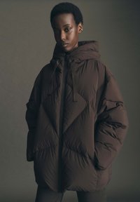 Giacca imbottita marrone con colletto alto, zip frontale e stringere. Presenta una texture trapuntata e una silhouette oversize.