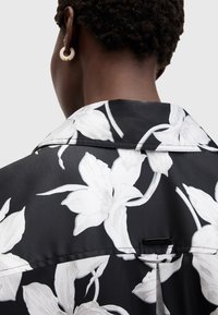 Personne avec les cheveux courts et bouclés portant une chemise noire avec de grandes impressions florales blanches et une petite étiquette noire rectangulaire dans le dos.
