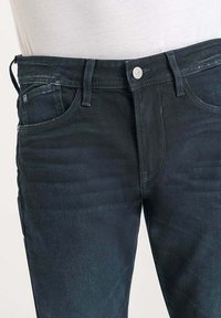 Jeans en denim bleu foncé avec une coupe ajustée, dotés de cinq poches, d'une fermeture à bouton sur le devant, et de légères marques de frottement sur le tissu.