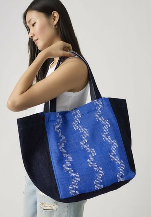 Donna con capelli lunghi e scuri che indossa una canottiera bianca e jeans strappati, tiene una grande borsa nera a tracolla con pannello frontale decorato da motivo blu.
