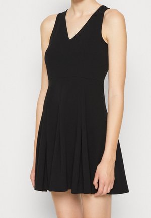 Kobieta w czarnej sukience bez rękawów z dekoltem w kształcie litery V i plisowanym dołem, stojąca na tle prostego, jasnego tła.