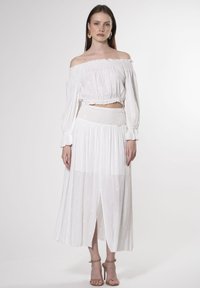 Hvid off-the-shoulder bluse med sammensamlet kant og lange ærmer, parret med en flydende hvid nederdel med elastisk talje og blomsterbroderi.