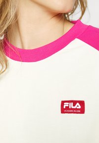 T-shirt en coton blanc avec col et manches côtelés roses. Présente un logo FILA rouge en patch sur la poitrine. Texture lisse avec un design décontracté.