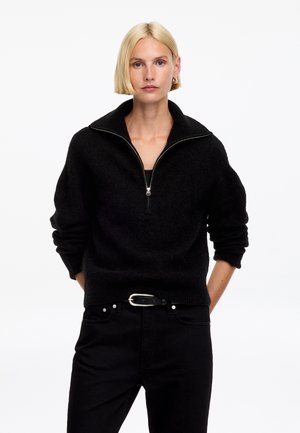 Mujer rubia con el pelo corto que lleva un suéter negro con cremallera y pantalones negros con cinturón, de pie frente a un fondo blanco liso.