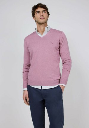 Hombre con suéter rosa claro con cuello en V sobre camisa blanca con cuello y pantalones azul marino, de pie frente a un fondo liso.