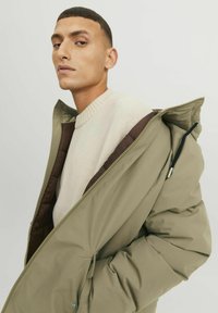 Veste matelassée vert olive avec fermeture éclair intégrale, dotée d'une doublure marron et d'une capuche avec cordon de serrage. Portée par-dessus un pull texturé crème.