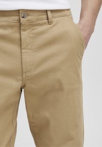 Chinos en coton beige à coupe droite, avec une fermeture à un bouton, des poches latérales et un tissu lisse et texturé.