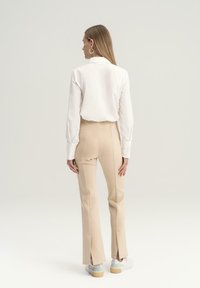 Camisa blanca de manga larga y pantalones beige de talón alto con aberturas laterales, combinados con zapatillas blancas que tienen un acento azul claro.