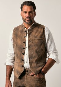 Chaleco sin mangas marrón hecho de un tejido texturizado, con botones negros, un pequeño bolsillo y un cuello alto, usado sobre una camisa blanca.