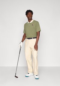 Zelena polo majica s kremastim ovratnikom i logom, uparena s bež hlačama. Dodaci uključuju golf palicu i bijele cipele s plavim akcentima.