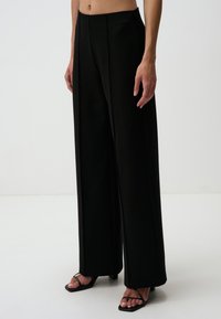 Pantalons noirs à jambes larges en tissu lisse, avec des plis à l'avant et une taille haute. Associés à des sandales noires à lanières.