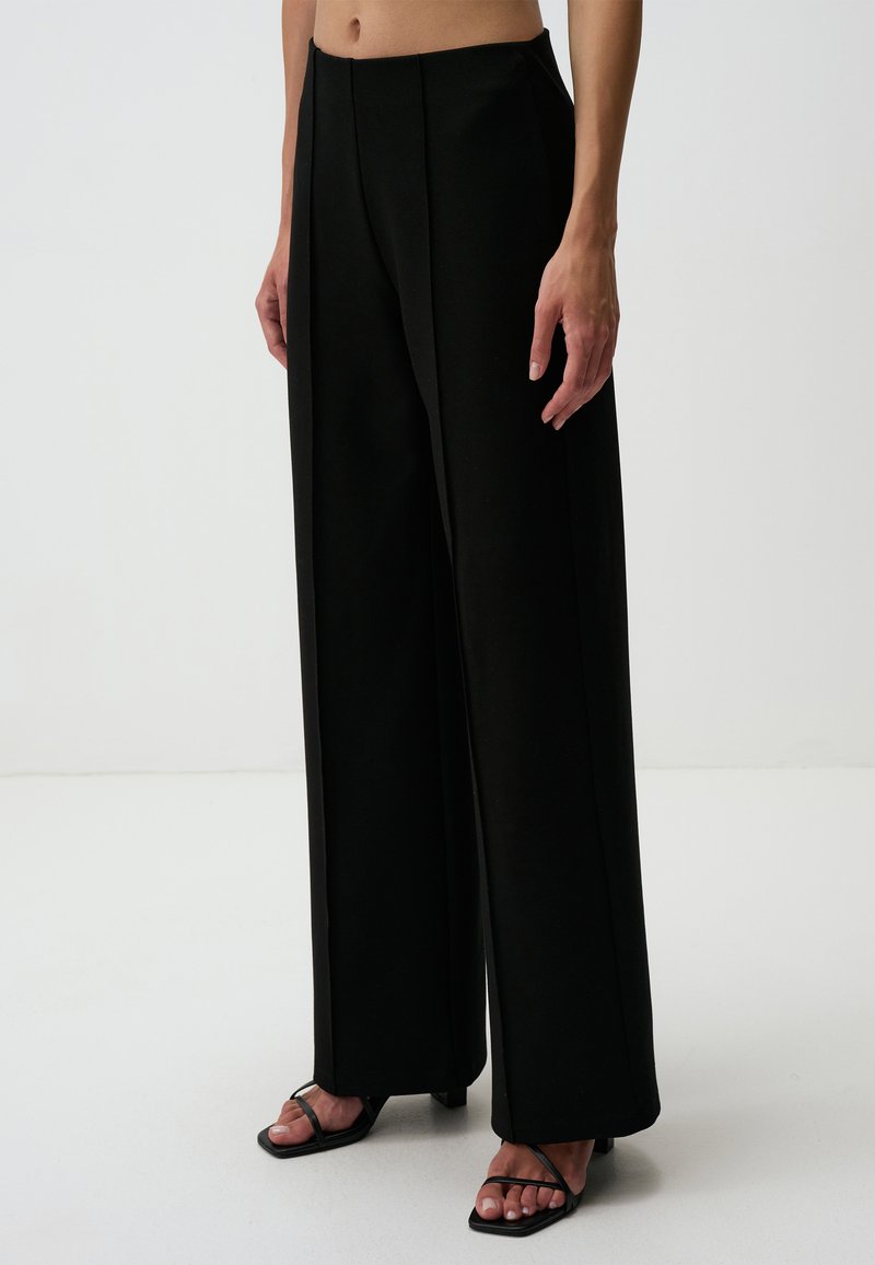 Pantalons noirs à jambes larges en tissu lisse, avec des plis à l'avant et une taille haute. Associés à des sandales noires à lanières.