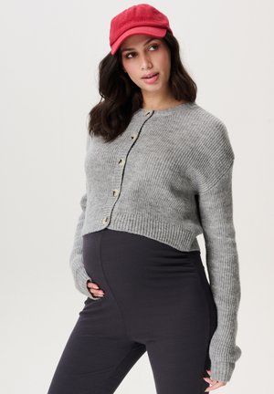 Grijze geribde cardigan met knopen, gedragen over donkere leggings. Model houdt haar zwangere buik vast en draagt een rode baseballcap.