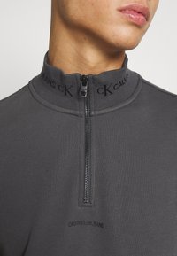 Gros plan sur une personne portant un haut zippé Calvin Klein gris foncé avec le logo sur le col et la poitrine, mettant en avant le cou et le bas du visage.