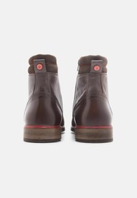 Botas de tobillo de cuero marrón con un cuello de ante marrón texturizado, costuras rojas y un acento de logo rojo redondo en la parte trasera. Suela plana y estratificada.