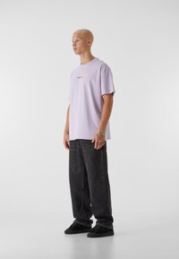 Lys lilla bomull t-skjorte med rund hals og korte ermer, kombinert med svarte baggy jeans og svarte joggesko. Enkel logo i midten.