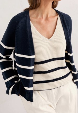 Femme portant un cardigan rayé bleu marine et blanc par-dessus un pull crème à col en V avec des rayures bleu marine, assorti à un pantalon blanc.