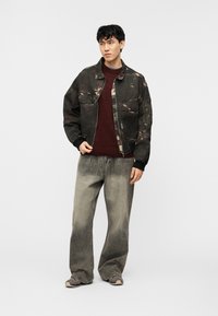 Νεαρός άνδρας που φοράει σκουρόχρωμο στρατιωτικό μπουφάν πάνω από μια βουργουνδίας μπλούζα, ξεθωριασμένα γκρι τζιν με φαρδιά μπατζάκια και γκρι αθλητικά παπούτσια, στέκεται μπροστά σε απλό φόντο.