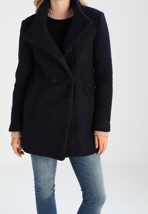 Frau trägt einen dunkelblauen, zweireihigen Wollmantel über einem schwarzen Oberteil mit rosa Strickkragen und hellblauen Jeans.