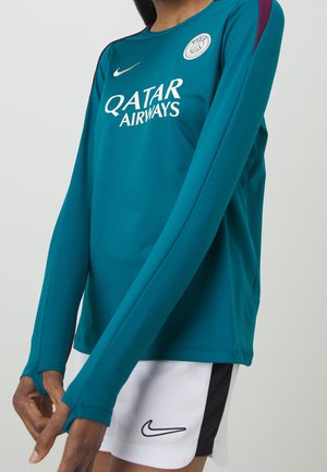 Persona vistiendo una camiseta de fútbol de manga larga color verde azulado del Paris Saint-Germain con el logo de Qatar Airways y pantalones cortos blancos de Nike con una franja negra en el lateral.