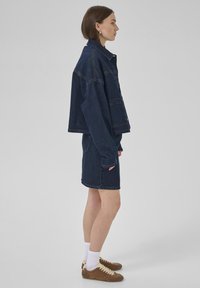 Veste en denim et jupe bleues foncées, avec une coupe décontractée, de grandes poches et des manches retroussées ; associées à des baskets en daim marron et des chaussettes blanches.
