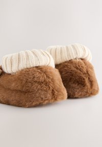 Chaussons en fausse fourrure marron avec un large revers tricoté crème, de forme arrondie et texture douce, offrant un design moelleux et confortable.
