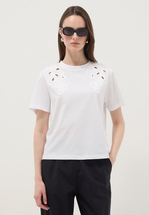 Vrouw draagt een wit T-shirt met korte mouwen met bloemenuitsnedes op de schouders, zwarte broek, zwarte zonnebril en gouden oorbellen, staand tegen een effen achtergrond.