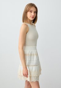 Un débardeur côtelé beige clair s'associe avec une jupe blanche en tissu texturé, ornée de dentelle et de motifs rayés horizontaux.