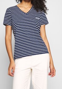 Kobieta ubrana w granatowo-biały T-shirt w poziome paski z dekoltem w serek i krótkim rękawem oraz białe spodnie, pokazana od ramion do ud.