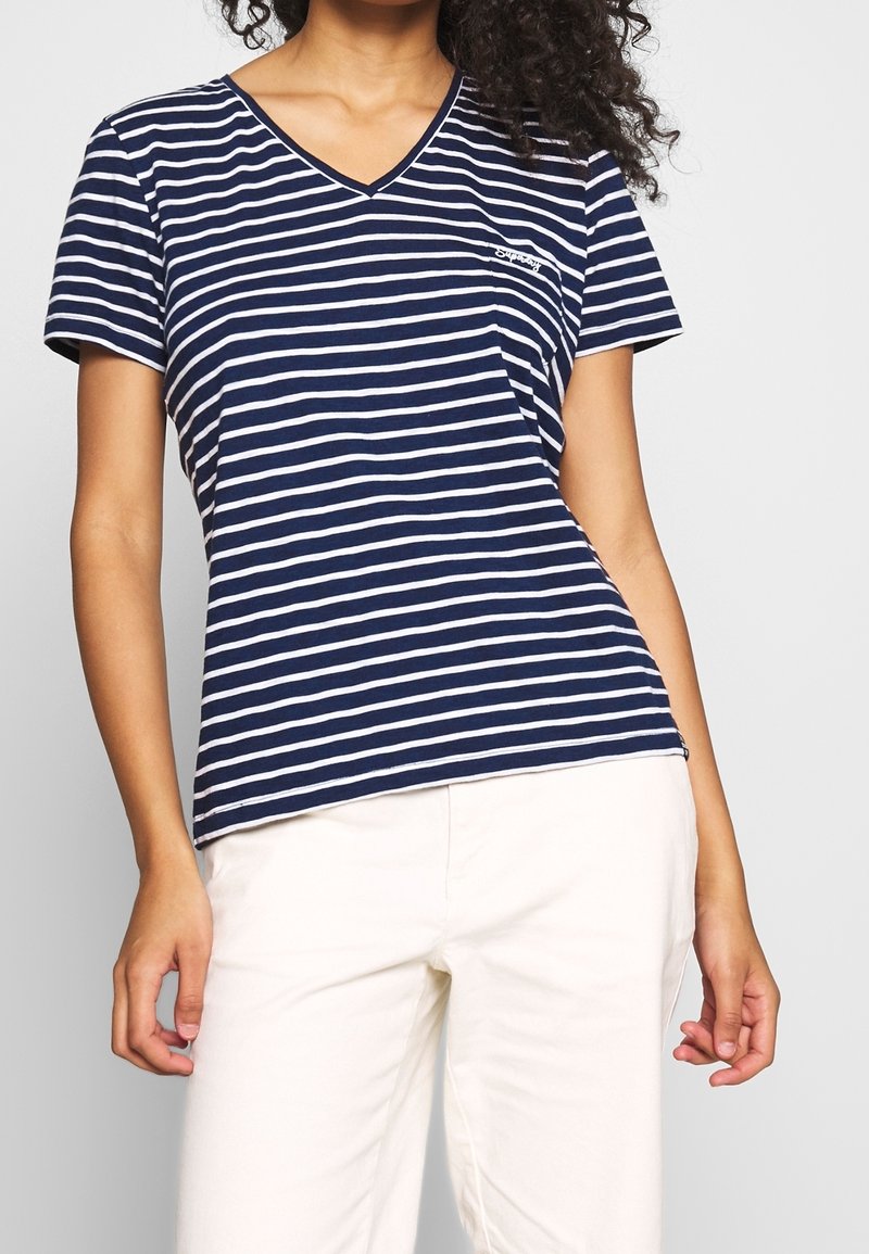 Kobieta ubrana w granatowo-biały T-shirt w poziome paski z dekoltem w serek i krótkim rękawem oraz białe spodnie, pokazana od ramion do ud.