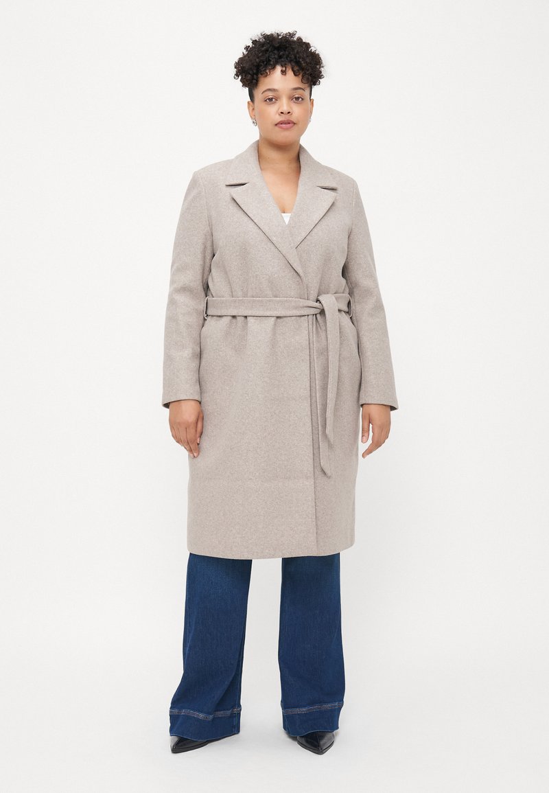 Cappotto beige con cintura, colletto a rever e maniche lunghe, realizzato in un tessuto morbido. Abbinato a pantaloni in denim blu a gamba larga e scarpe nere.