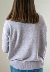 Persoon met schouderlang bruin haar, gekleed in een lichtgrijze trui en blauwe spijkerbroek, met de rug naar de camera toe tegen een effen achtergrond.