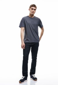 Graues Baumwoll-T-Shirt mit Rundhalsausschnitt und dezentem "BRUSSELS"-Text, kombiniert mit dunkelblauen Jeans und schwarzen Sneakers mit weißen Schnürsenkeln.