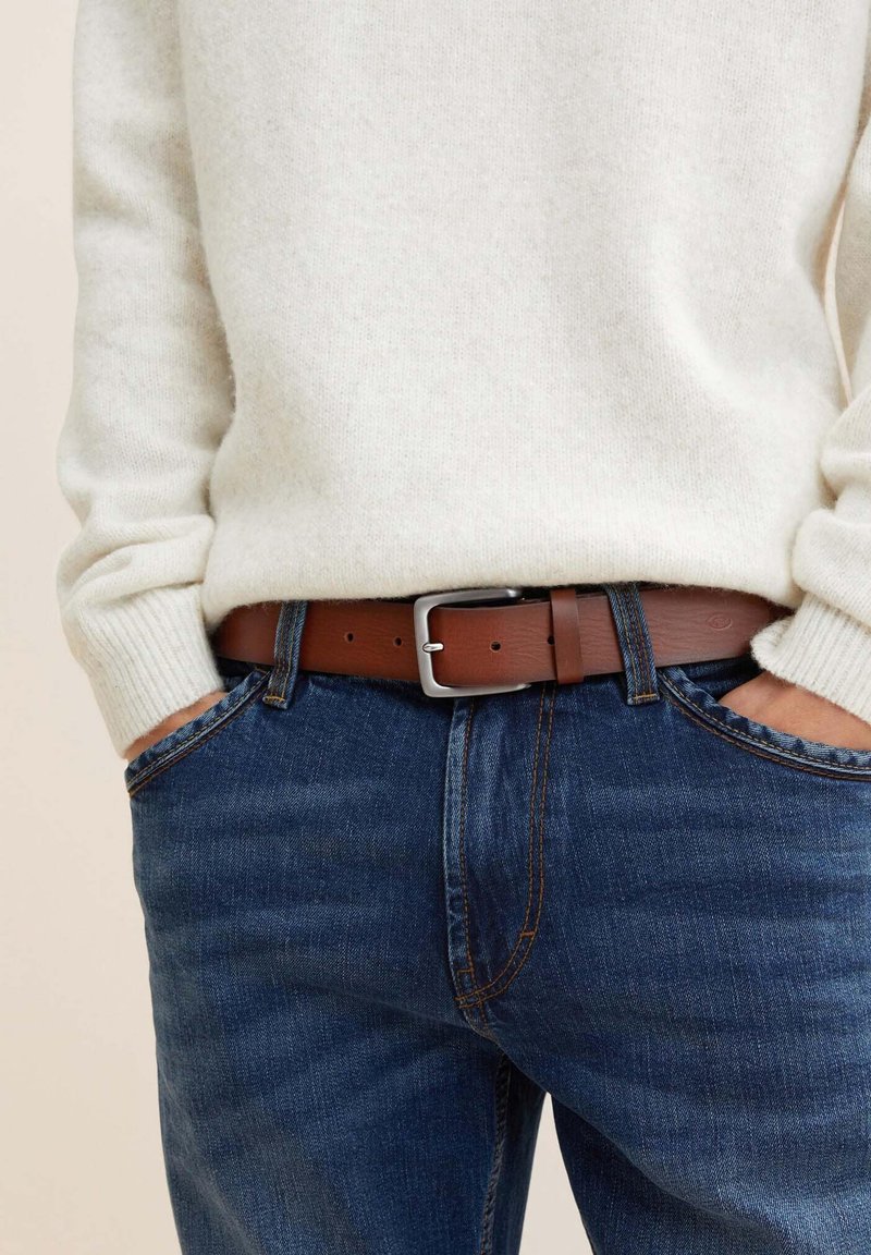 Ceinture en cuir marron avec boucle en argent, portée avec un pull crème et un jean bleu foncé, montrant une texture douce et un style décontracté.