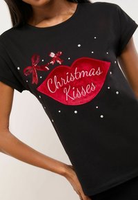 Melna īsām piedurknēm T-kreklā, kas rotāts ar teksturētu sarkano lūpu grafiku, uzrakstu "Ziemassvētku skūpsti" un sekvīnu loku, rotātiem ar baltām pērlītēm.