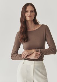 Frau mit langen braunen Haaren, die ein braunes Langarmoberteil und weiße hochtaillierte Hosen trägt, mit vor der Taille verschränkten Armen vor einem schlichten Hintergrund.