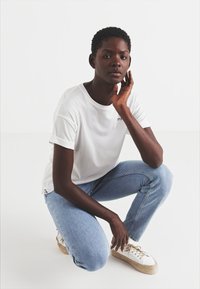 Hvitt kortermet t-skjorte, blå jeans og hvite sneakers med strådetaljer. Modellen poserer i en krum stilling foran en ensfarget bakgrunn.